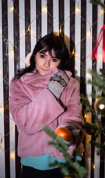 Russian 48 years old woman in pink coat holding tangerines against yellow christmas lights before Christmas