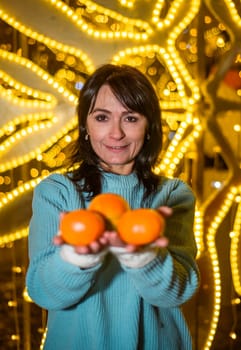 Russian 48 years old woman in blue sweater holding tangerines against yellow christmas lights before Christmas