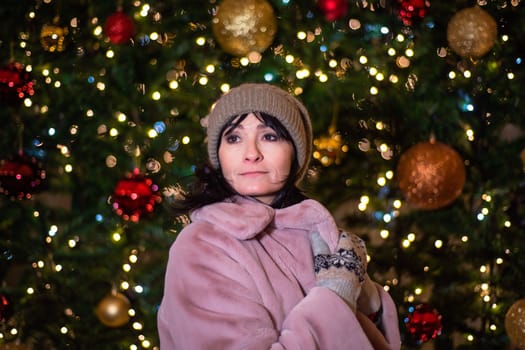 Russian 48 years old woman in pink coat standing against christmas tree and lights before New Year