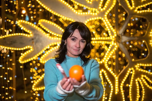 Russian 48 years old woman in blue sweater holding tangerines against yellow christmas lights before Christmas
