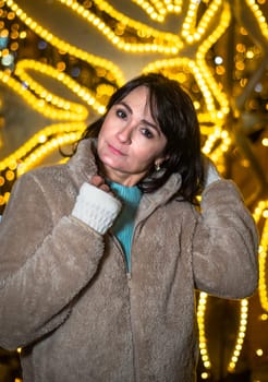 Russian 48 years old woman in blue sweater and brown coat standing against yellow christmas lights before Christmas