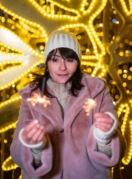 Russian 48 years old woman in blue sweater and pink coat standing against yellow christmas lights before Christmas and holding lit sparklers