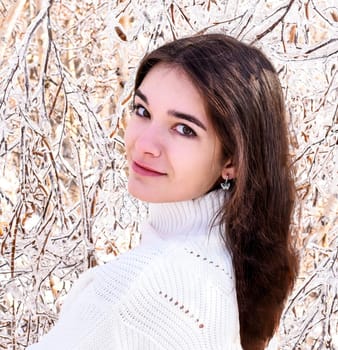 Portrait of young Russian woman in white sweater looking at camera next to frozen branches of birch trees, covered with ice. Winter forest in Russia