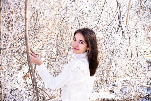 Portrait of young Russian woman in white sweater looking at camera next to frozen branches of birch trees, covered with ice. Winter forest in Russia