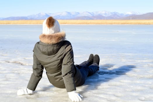 Young woman in warm clothes sitting on the ice and looking at snowy mountains. winter, Russia