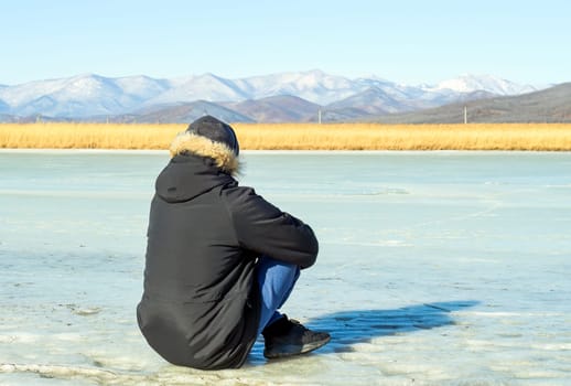 Young man in warm clothes sitting on the ice and looking at snowy mountains. winter, Russia.