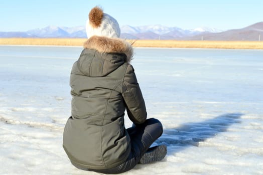 Young woman in warm clothes sitting on the ice and looking at snowy mountains. winter, Russia
