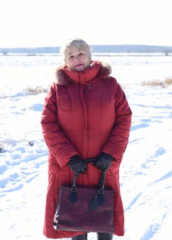 Russian 67 years old senior woman in red coat looking at the camera against snowy field. winter