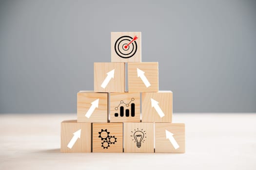 Cube wood blocks arranged with Target icon on top rise up arrows, representing growth. Bar graph chart steps on vertical symbolize a business growth process, signifying profit, investment, economic