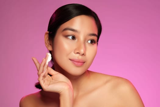 A young woman with smooth skin applies a skincare product to her face, showcasing a natural look and soft lighting in the background during evening hours.