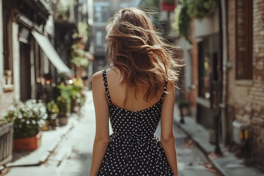 Back view of young woman in stylish black dress with white polka dots on city street on summer day.
