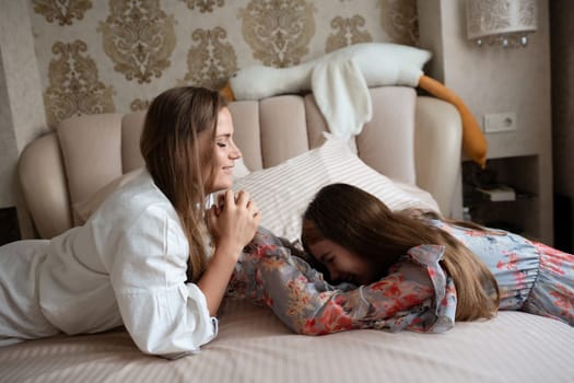 Two women, one in a white robe and one in a patterned dress, lie on a bed. They're close, one leaning toward the other affectionately. Intimate moment of shared closeness.