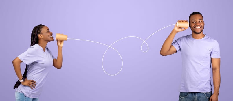 Communication and Fun. Portrait of happy casual black couple talking through tin can telephone isolated on violet studio background. Cheerful boyfriend and girlfriend hearing and speaking through cups