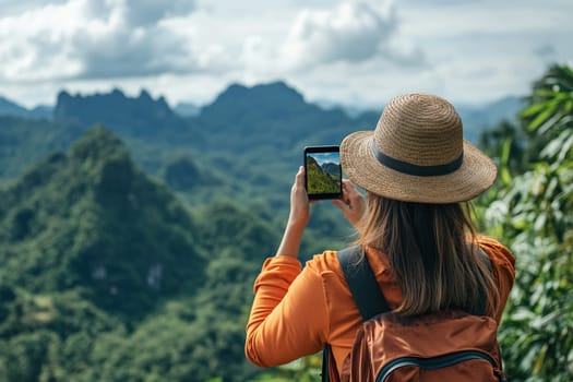 A female tourist with a backpack on her back takes a photo of a mountain landscape on her smartphone while standing on top of a cliff.