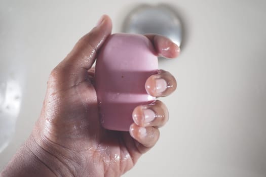 Hands are holding a pink soap bar under running water in a bathroom.