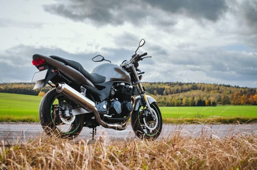 A gray Motorcycle stands by the road. The road is empty, autumn landscape, bright green field, yellow dry grass, cloudy sky. Traveling by motorcycle