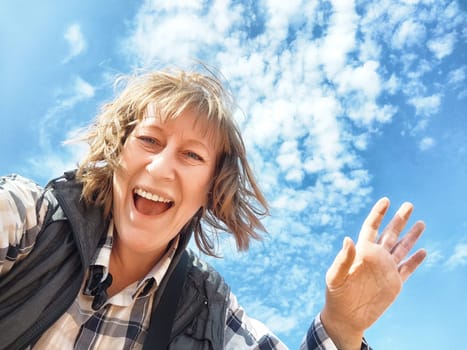 Middle-aged woman enjoys a cheerful moment while taking a selfie against a backdrop of fluffy clouds and blue sky.
