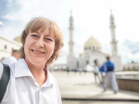 A middle-aged blonde woman smiles for a selfie while exploring a stunning mosque during her travels.