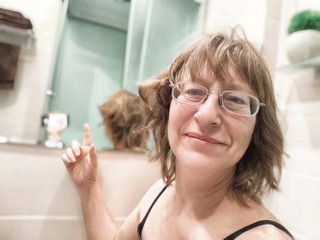 A middle-aged woman takes a morning selfie in her bathroom mirror, smiling and enjoying her blogging journey.