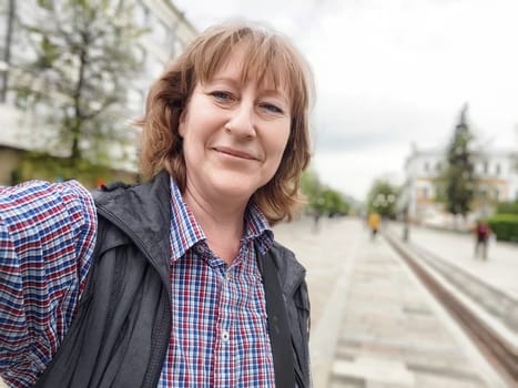 A middle-aged female traveler captures a cheerful selfie while exploring a lively cityscape on a beautiful day.