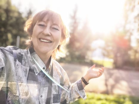 A middle-aged female blogger takes a cheerful selfie in a sunlit outdoor setting surrounded by nature.