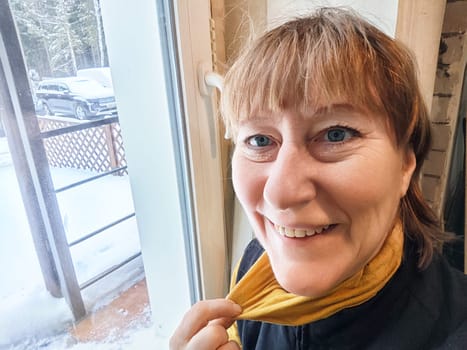 A middle-aged blonde woman captures a cheerful selfie indoors, surrounded by a snowy landscape and trees.