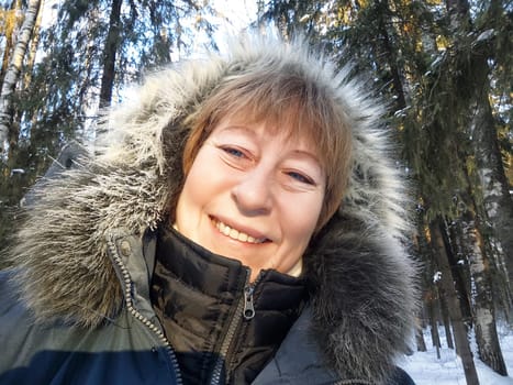 Mature blonde woman smiles while capturing a selfie in a beautiful snowy forest during her winter trip.