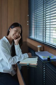 Young woman smiling while sitting at a desk with notepad, coffee, and natural light streaming through blinds.