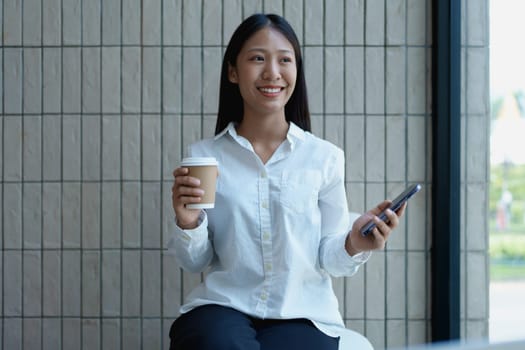 Young woman sitting in a modern cafe, holding a coffee cup and smartphone, smiling and enjoying her time indoors.