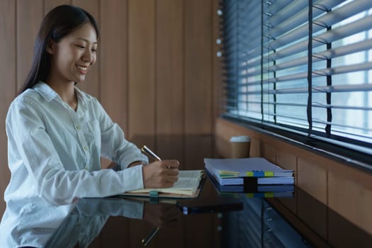 Young woman writing in notebook at modern desk with natural light streaming through blinds in contemporary office space.