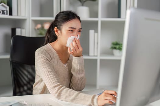 A young woman wipes her nose with a tissue, looking at her computer, indicating she is feeling unwell in a professional environment.