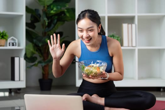 A cheerful woman waves at her laptop, holding a bowl of salad, representing a healthy lifestyle and social connection.