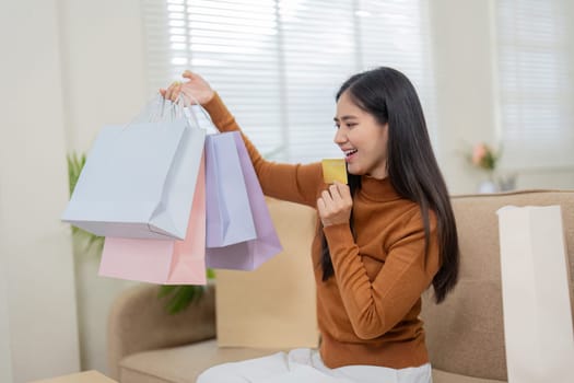A cheerful woman holds her credit card and shopping bags, reveling in her online shopping achievements at home.