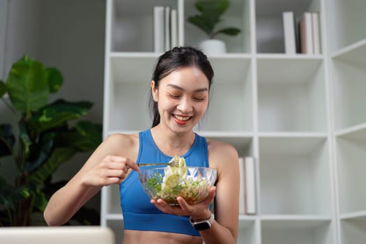 A joyful woman in athletic wear happily eats a salad while engaging with a laptop, emphasizing health and nutrition.