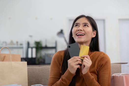 A cheerful woman holds two credit cards and a smartphone, contemplating her online shopping decisions at home.