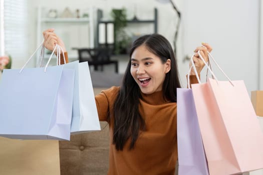 A joyful young woman holds colorful shopping bags while smiling at her smartphone, celebrating her online shopping experience at home.