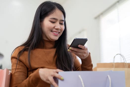A young woman smiles as she checks her online shopping on her smartphone, surrounded by colorful shopping bags at home.