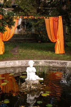 fountain in the form of an angel sitting in water, 19th century. Behind is a photo zone made of draped orange curtains. Around are green trees.water lilies grow in the fountain.