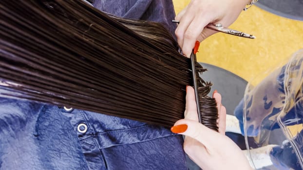 In the bustling atmosphere of a salon, a little girl sits patiently as a stylist with polished red nails carefully trims her damp hair. The sheen of the scissors and the precise hand movements capture the delicate process of styling a child's hair.