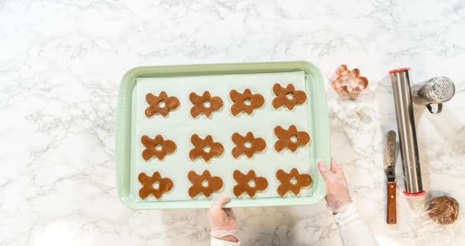 Angled view of a baker's hands lifting a baking tray with gingerbread man-shaped cookies ready to go into the oven, set against a marble countertop with tools visible.