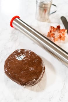 A ball of Chocolate Ginger Sugar Cookie dough wrapped in plastic, placed on a marble surface with baking tools in the background. Close-up perspective.
