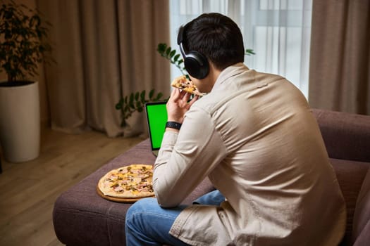 Young man sitting on sofa and eating pizza while watching movie on laptop