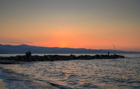 As the sun sets behind distant mountains, a lone figure enjoys the serene beauty of the peaceful shoreline.
