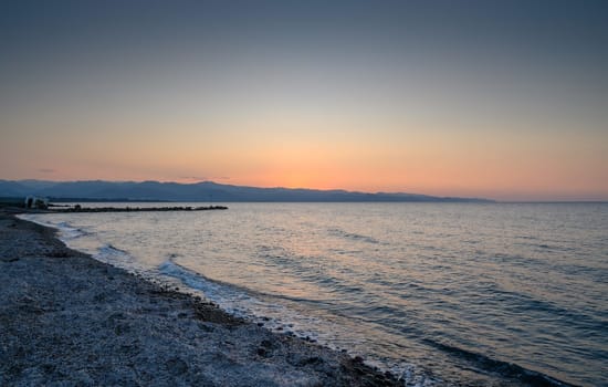 Waves gently lap against the shore as the sun sets, casting a warm glow on the distant mountains and calm sea.