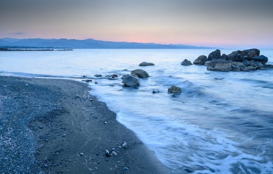 Gentle waves lap against pebbles as the sun rises, casting warm colors over the serene coastal landscape.