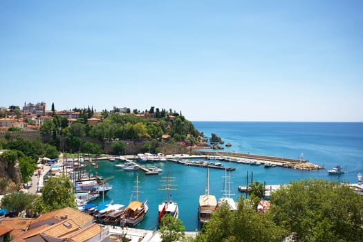 A panoramic view of Antalya, Turkey 09.20.2024 harbor, featuring traditional wooden boats, turquoise waters, historic architecture, lush greenery, and a peacefulc oastline under a clear sky.