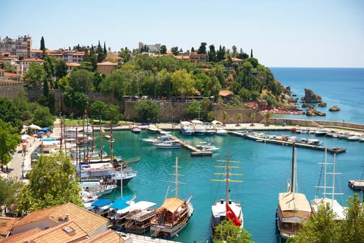A stunning view of Antalya marina, featuring traditional wooden boats, turquoise waters, historic stone walls, lush greenery, and a scenic coastal backdrop under a sunny sky.