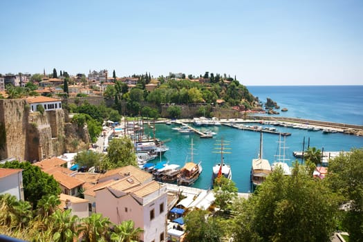 A stunning panoramic view of Antalya marina, showcasing traditional sailboats, turquoise waters, ancient stone walls, and charming Mediterranean-style houses surrounded by lush greenery.