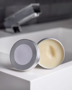 Opened body butter jar with blank label on round lid on washbasin near modern faucet in dark grey masculine bathroom close up, mockup. Natural herbal unbranded cosmetics for bodycare and spa