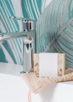 Beige handmade soap bar with blank label on sisal soap saver bag on washbasin near faucet in green tropical bathroom close up, mockup, copy space. Natural herbal unbranded product for daily hygiene